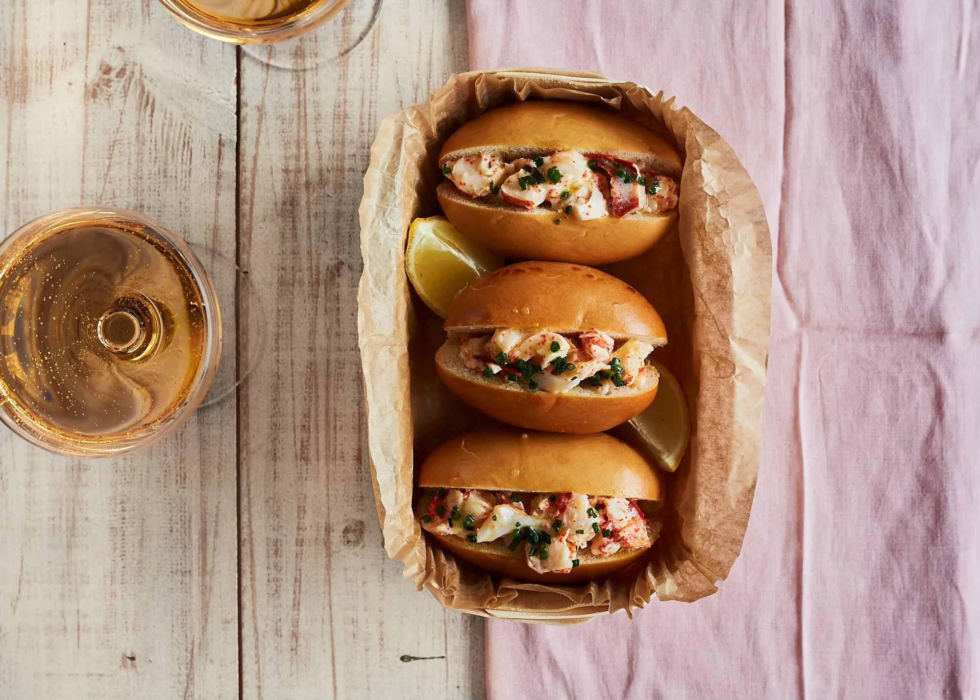 Lobster rolls in a paper-lined basket with lemon and two glasses of sparkling wine